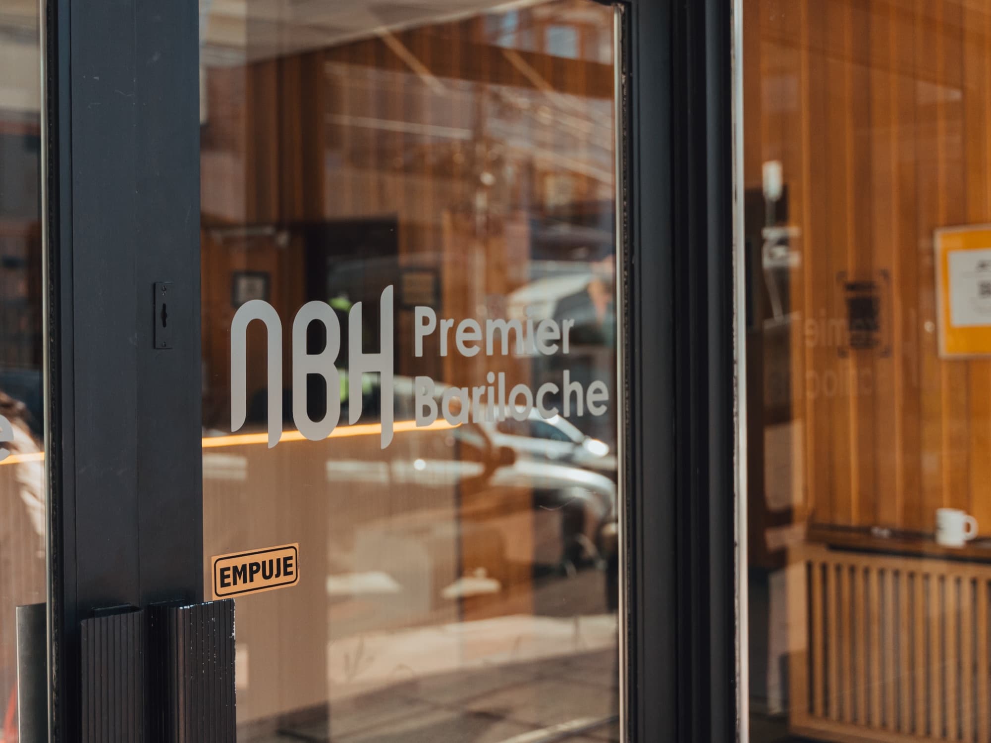 Puerta de entrada de NBH Premier Bariloche, con el logo del hotel sobre el vidrio y la recepción de madera al fondo